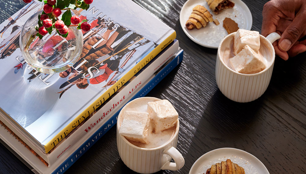 mugs of hot cocoa, pastries, a stack of books, and vase of holly